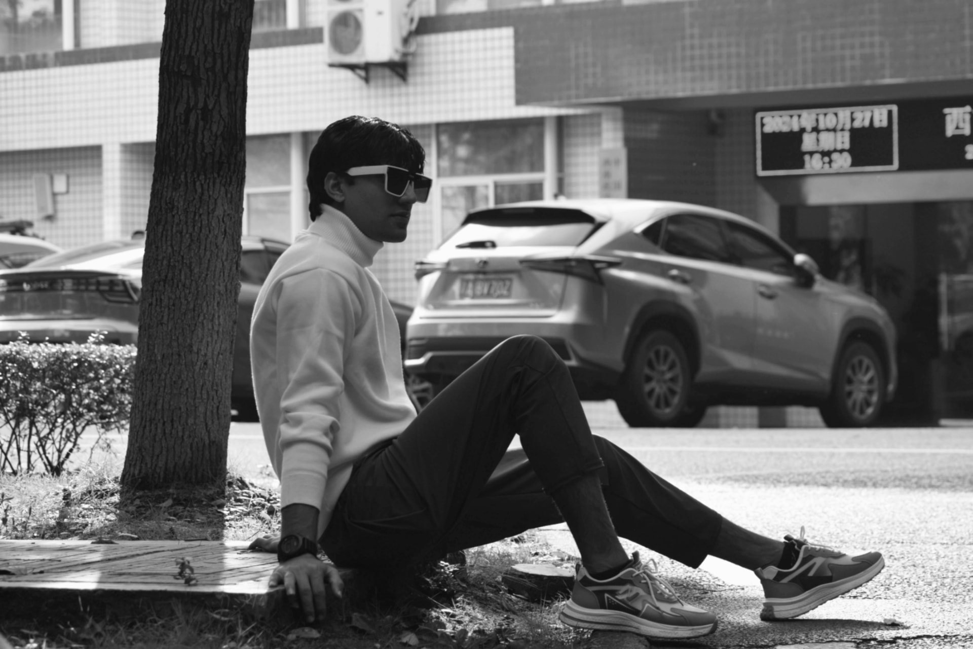 Black and white photo of a man in a white turtleneck and sunglasses sitting by a tree on a city street