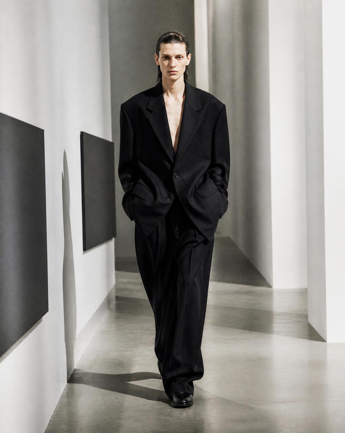 Model in oversized black tailoring walking through a white gallery corridor